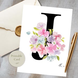 May include: A white greeting card featuring a large black letter J decorated with a watercolor floral arrangement in shades of pink, purple, and light blue. The card is positioned next to a gold pen and a cream envelope with a gold wax seal.