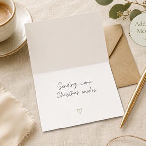 Pu&ograve; includere: Un biglietto d'auguri bianco aperto con il testo stampato in nero Sending warm Christmas wishes e una piccola icona a forma di cuore in basso. Il biglietto &egrave; posizionato accanto a una busta marrone, una penna dorata e caff&egrave; su tessuto chiaro.