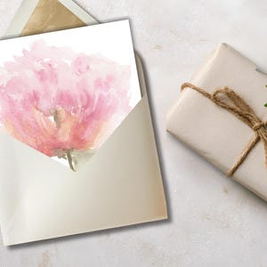 Printable watercolor pink peony card being tucked into a gold-lined envelope, styled next to a rustic brown paper wrapped gift with twine and a green leaf on a stone surface.