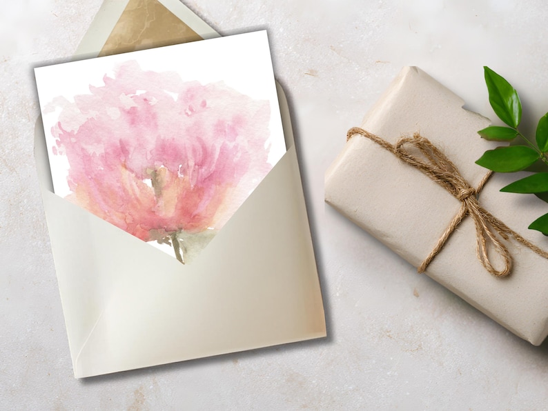 Printable watercolor pink peony card being tucked into a gold-lined envelope, styled next to a rustic brown paper wrapped gift with twine and a green leaf on a stone surface.