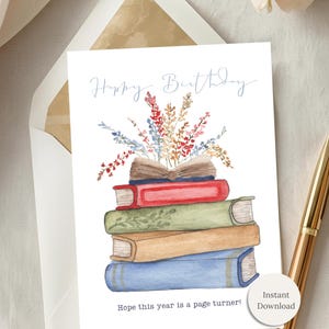 May include: A watercolor birthday card with a stack of books and floral accents. The card reads "Happy Birthday" and "Hope this year is a page turner!" A gold pen rests beside the card.