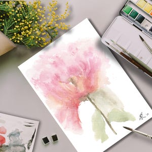 Behind-the-scenes view of a hand-painted watercolor pink peony surrounded by professional watercolor pans, brushes, and botanical yellow mimosa flowers on a studio workspace.