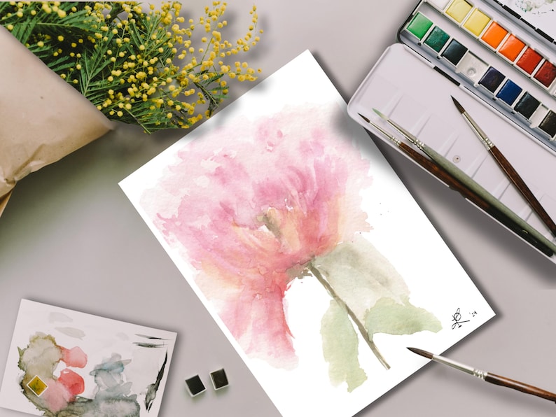Behind-the-scenes view of a hand-painted watercolor pink peony surrounded by professional watercolor pans, brushes, and botanical yellow mimosa flowers on a studio workspace.