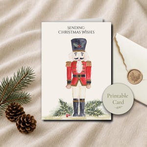 May include: A greeting card featuring a watercolor illustration of a nutcracker in a red uniform with a black hat, standing amidst green pine branches. The text reads SENDING CHRISTMAS WISHES. A circular label says Printable Card.