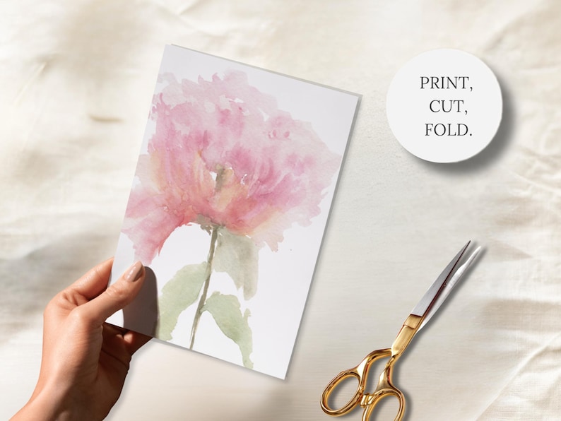 A hand holding a hand-painted card featuring a watercolor peony, gold scissors and a badge with the wording Print, Cut, Fold