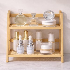 May include: A two-tiered bamboo shelf displays various beauty products. The top shelf holds a round perfume bottle, a rectangular perfume bottle, and a compact. The bottom shelf features serum bottles, and jars of powder and cream.