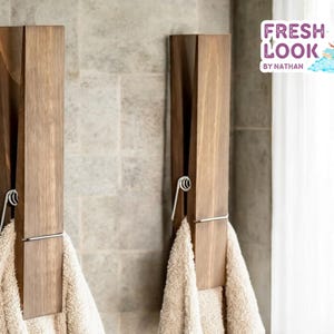 Può includere: Due ganci portasciugamani in legno, a forma di mollette giganti, sono fissati a una parete piastrellata. Ognuno ha una clip a molla in metallo e tiene un asciugamano beige. L'immagine include il testo "FRESH LOOK by NATHAN".