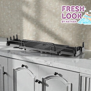 Puede incluir: Una bandeja de bañera negra y extensible con un soporte para libros o tabletas incorporado. La bandeja es de metal y se encuentra sobre una encimera blanca. La imagen incluye el texto "FRESH LOOK by NATHAN".