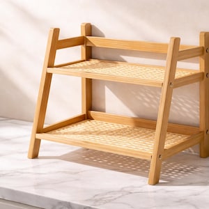 May include: A two-tiered, light brown bamboo shelf with woven rattan shelves. The shelf has a rectangular shape with angled side supports and is designed for storage or display. The shelf is placed on a white marble surface.