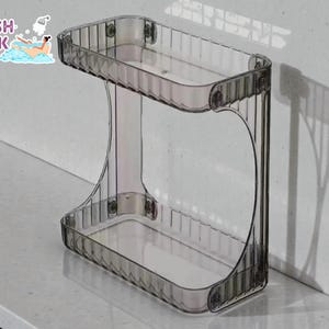 Könnte beinhalten: Ein klares, zweistufiges Regal mit rechteckigem Design. Das Regal besteht aus transparentem Kunststoff und hat einen Grauton. Das Logo "FRESH LOOK BY NATHAN" befindet sich in der oberen linken Ecke. Das Regal ist für die Badezimmer- oder Küchenaufbewahrung konzipiert.