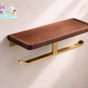 May include: A wooden shelf with a dark brown finish and a gold-coloured metal toilet paper holder. The shelf has a raised edge and is mounted on a wall. The text "FRESH LOOK BY NATHAN" is in the upper left corner.