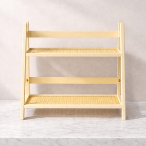 May include: A light-colored, two-tiered wooden shelf with woven rattan shelves. The shelf has a rectangular shape and is supported by four legs. The shelves are ideal for displaying decorative items or organizing small objects.