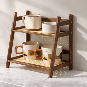 May include: A two-tiered wooden shelf with woven rattan shelves holds four ceramic mugs. The mugs are white, beige, and brown, with various designs. The shelf is dark brown and has an A-frame design.
