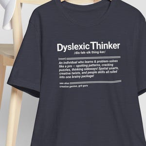 Puede incluir: Una camiseta azul marino con el texto "Dyslexic Thinker" en blanco. La camiseta incluye una definición de un pensador disléxico, destacando las habilidades de resolución de problemas y el pensamiento creativo. Una gorra de béisbol blanca descansa sobre una silla de madera.