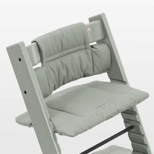 Puede incluir: Una trona de madera en color verde salvia claro con coj&iacute;n de asiento y respaldo acolchados a juego. La silla tiene una estructura con listones horizontales y una peque&ntilde;a etiqueta con logotipo visible en la parte superior derecha del respaldo.