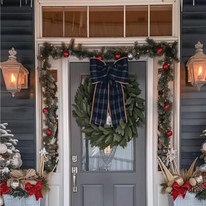 Puede incluir: Una puerta de entrada decorada para Navidad con una corona verde, un lazo a cuadros y guirnalda con adornos rojos. La puerta es gris y la casa tiene revestimiento azul. Dos faroles iluminados flanquean la puerta.