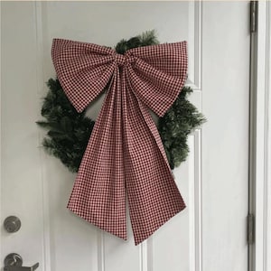 Puede incluir: Una corona navide&ntilde;a con un gran lazo a cuadros rojos y blancos. El lazo es el punto focal, con largas colas que fluyen. La corona est&aacute; hecha de ramas de pino artificiales verdes, creando una decoraci&oacute;n festiva.