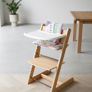 Puede incluir: Una trona de madera de color claro con una bandeja de pl&aacute;stico blanca, un coj&iacute;n para el asiento y otro para el respaldo. Los cojines presentan un estampado abstracto multicolor con tonos morados, verdes y rosas. La silla tiene niveles ajustables.