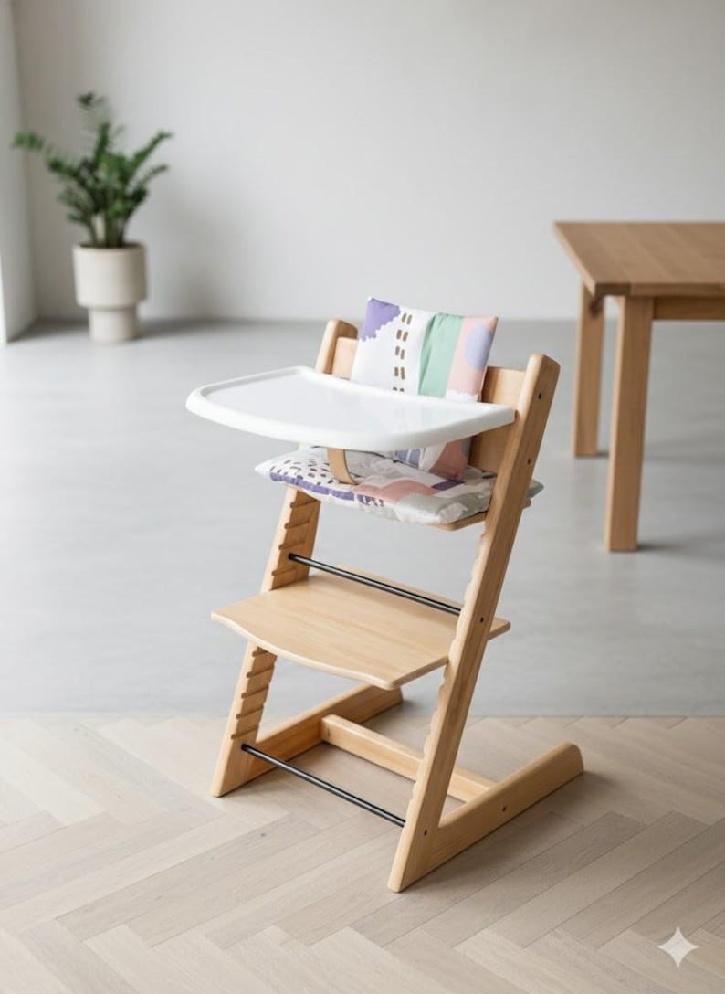 Puede incluir: Una trona de madera de color claro con una bandeja de pl&aacute;stico blanca, un coj&iacute;n para el asiento y otro para el respaldo. Los cojines presentan un estampado abstracto multicolor con tonos morados, verdes y rosas. La silla tiene niveles ajustables.