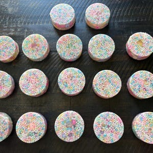 May include: Fifteen round, colorful bath bombs are arranged on a dark wooden surface. Each bath bomb is covered in small, pastel-colored beads. The bath bombs have a pink base and a speckled appearance.