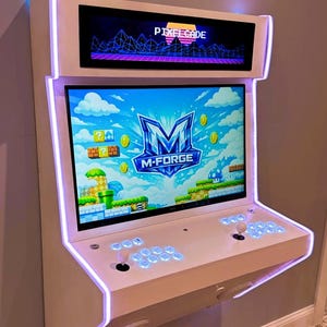May include: A white arcade machine with a large screen displaying a video game. The machine has a retro design with a top screen displaying "Pixelcade". The main screen shows "M-FORGE". It features joysticks and buttons.