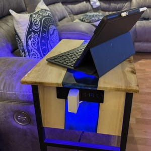 May include: A wooden side table with a black metal frame. The tabletop is light-colored wood with a blue resin inlay. A black tablet with a keyboard and stylus sits on the table. A white charger is plugged into the side.