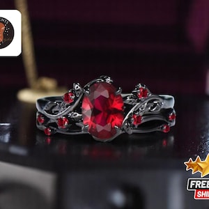 Może przedstawiać: Czarny pierścionek z dużym, owalnym, czerwonym kamieniem otoczonym mniejszymi czerwonymi kamieniami. Pierścionek ma wzór przypominający winorośl.