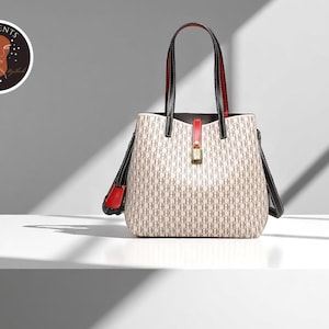 May include: A beige tote bag with a repeating pattern, black handles, and red accents. The bag has a gold-colored lock and a red strap. The bag is sitting on a white surface.