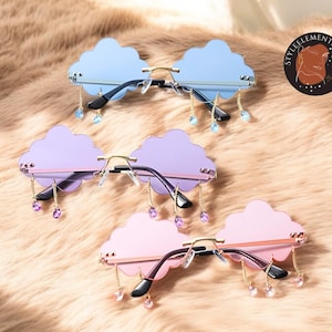 Puede incluir: Tres pares de gafas de sol en forma de nube con colgantes de cristal. Las gafas de sol son de color azul, morado y rosa.