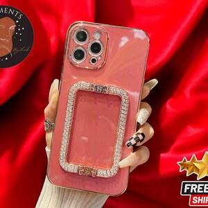 May include: A pink phone case with a gold frame and rhinestones. The case has a cutout for the camera lens and a decorative ring holder.