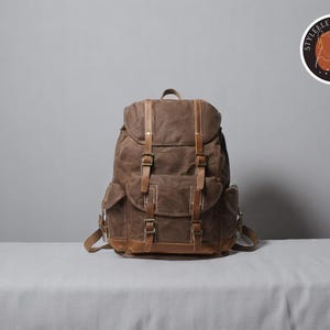 Puede incluir: Una mochila de lona marrón con correas y detalles de cuero. La mochila tiene múltiples bolsillos y un cierre de solapa. La bolsa está diseñada para actividades al aire libre y viajes. La bolsa tiene un estilo vintage.