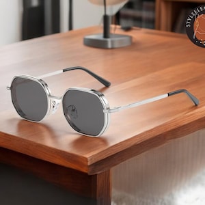 May include: Silver-framed sunglasses with dark grey lenses. The sunglasses have a geometric shape and are resting on a wooden table.