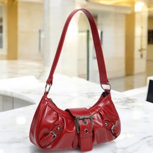 May include: A red leather handbag with a buckle closure and a single strap. The bag has a slightly curved shape and a smooth, shiny finish.