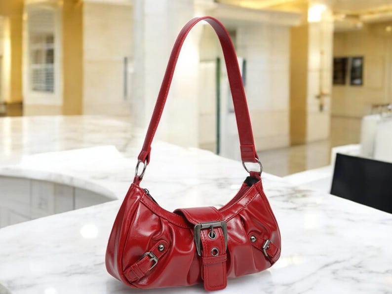 May include: A red leather handbag with a buckle closure and a single strap. The bag has a slightly curved shape and a smooth, shiny finish.