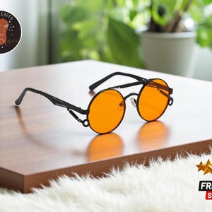 Könnte beinhalten: Schwarze runde Sonnenbrille mit orangenen Gläsern und einem Metallrahmen. Die Sonnenbrille hat ein einzigartiges Design mit einer Metallstange, die die Gläser verbindet.