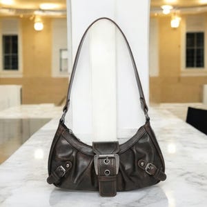 May include: A brown leather handbag with a silver buckle and a short strap. The bag has a curved shape and a slightly distressed look.