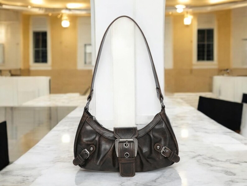 May include: A brown leather handbag with a silver buckle and a short strap. The bag has a curved shape and a slightly distressed look.