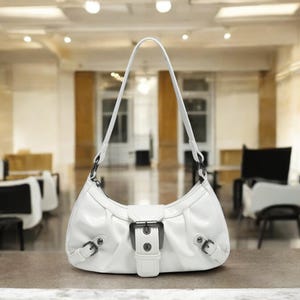 May include: A white leather handbag with a silver buckle and a short strap. The bag is sitting on a white surface.