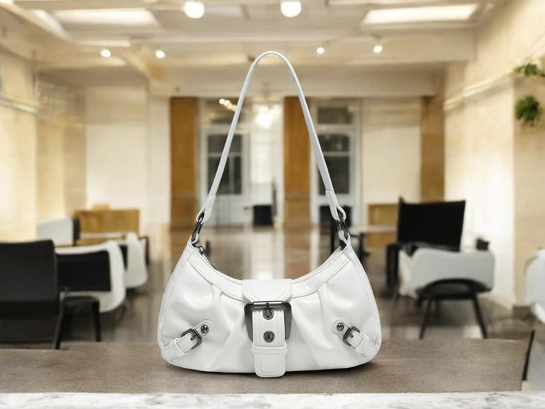 May include: A white leather handbag with a silver buckle and a short strap. The bag is sitting on a white surface.