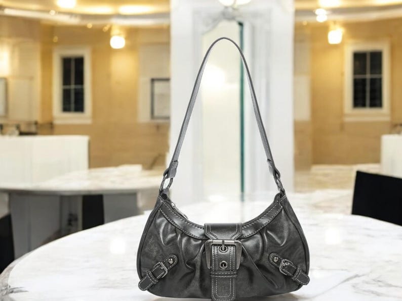 May include: A gray leather handbag with a single strap and a buckle closure. The bag has a slightly curved shape and a soft, slouchy look.