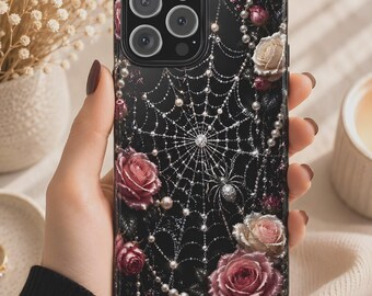 May include: Black phone case featuring a spiderweb design with pearls and roses. The case has a glossy finish and a raised texture. The roses are in shades of pink and cream. The phone case is held in a hand.