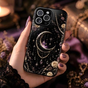 May include: Black phone case featuring a celestial design with a crescent moon, stars, and floral accents. The case has a glossy finish and is adorned with gold and purple details. The phone case is held in a hand with purple nail polish.