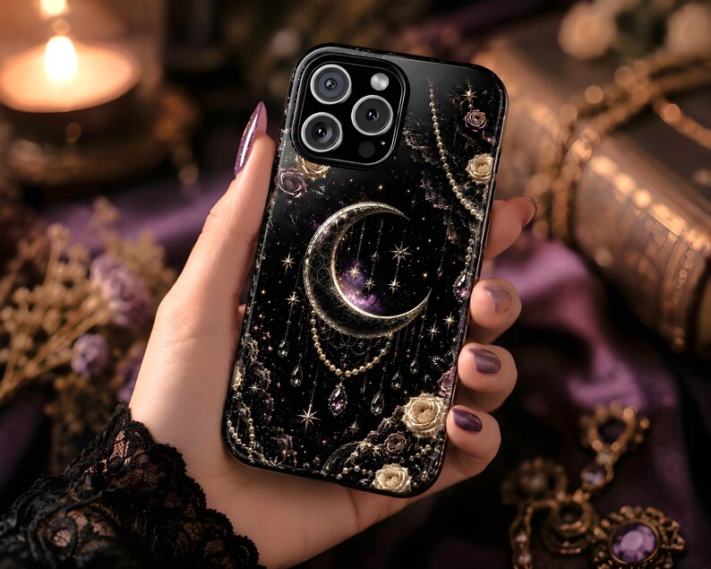 May include: Black phone case featuring a celestial design with a crescent moon, stars, and floral accents. The case has a glossy finish and is adorned with gold and purple details. The phone case is held in a hand with purple nail polish.