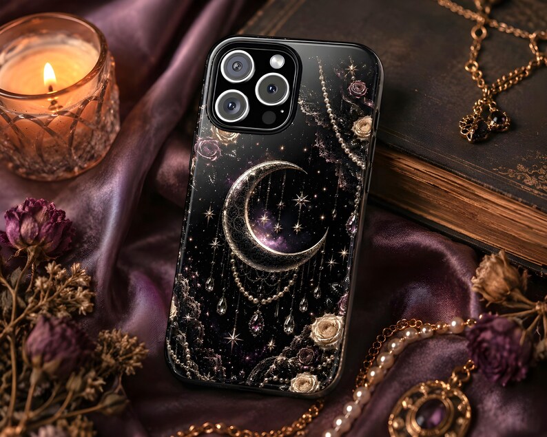 May include: Black phone case with a celestial design featuring a crescent moon, stars, and beaded accents. The case has a glossy finish and is surrounded by gold-colored jewelry and dried flowers. A lit candle sits in a glass holder.