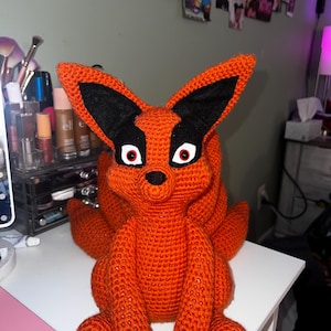May include: A crocheted orange fox plush toy with black ears and red eyes. The fox is sitting on a white surface.