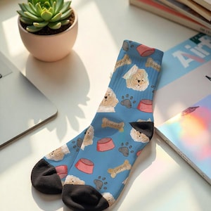 Puede incluir: Un par de calcetines azules con un estampado repetido de perros blancos, cuencos rosas, huellas de patas y galletas con forma de hueso. Los calcetines tienen talones y punteras negras. La palabra "Biscuit" está impresa en las galletas.