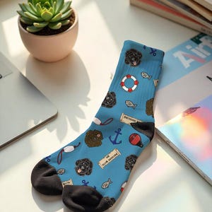 May include: A pair of blue socks with a pattern of black dog faces, anchors, fish, life preservers, and the text "Portuguese Water". The socks have black heels and toes. A succulent plant in a pink pot is in the background.