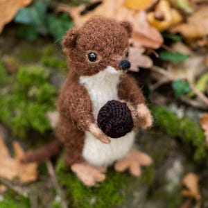 Op de afbeelding: Een handgemaakte, miniatuur bruin-witte wezel die een donkerbruine bal vasthoudt. De wezel heeft een zachte textuur en staat tegen een achtergrond van herfstbladeren en met mos bedekte rotsen. Een charmant bosdier.