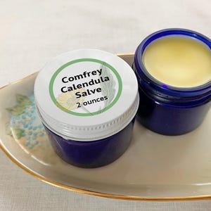 May include: Two cobalt blue glass jars containing a pale yellow salve. One jar is closed with a white lid featuring a green-bordered label that reads Comfrey Calendula Salve 2 ounces. The other jar is open, revealing the smooth yellow balm inside.