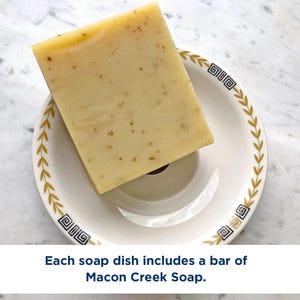 May include: A rectangular bar of light yellow soap with visible brown flecks resting on a white ceramic dish. The dish features a decorative gold leaf pattern and black geometric accents. The text reads: Each soap dish includes a bar of Macon Creek Soap.